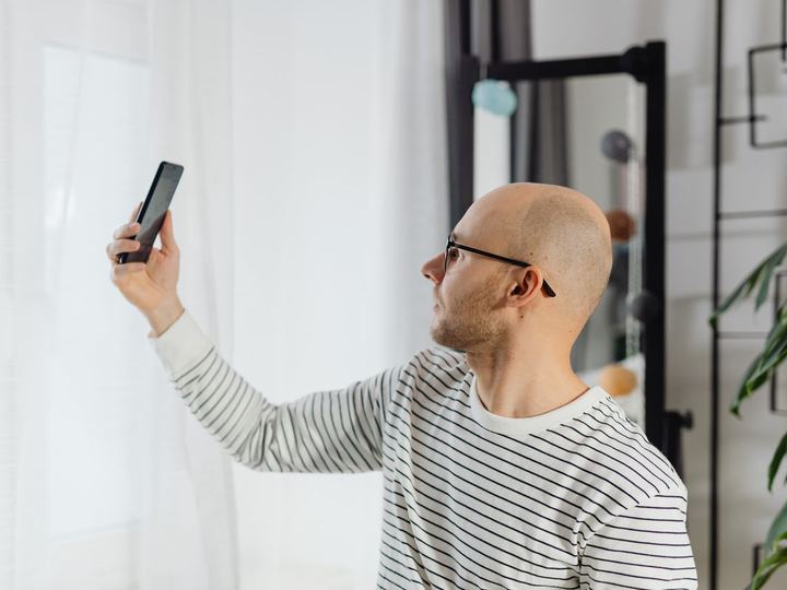 Hombre con calvicie revisándose la cabeza con el celular frente al espejo